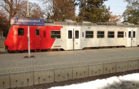 Bahnhof Langenzersdorf, © Leader-Region Weinviertel / Lahofer Ein Zug steht am Bahnhof Langenzersdorf, mit einem Schild im Vordergrund. Der Zug ist rot-weiß und die Umgebung ist von Bäumen umgeben.