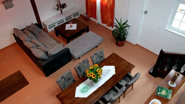 Blick von Stiegenaufgang im Loft 1, © Carlos de Mello Blick von oben auf ein Wohnzimmer mit grauem Sofa, Esstisch mit Sonnenblumen, roten Vorhängen und einer Pflanze.