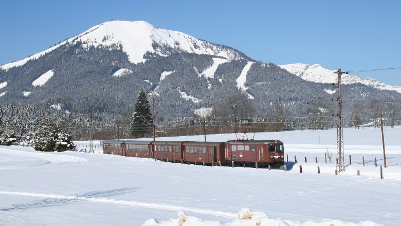 Familien-Erlebniszug Ötscherbär, © PROKOP Ein Zug fährt durch eine verschneite Landschaft mit einem schneebedeckten Berg im Hintergrund.