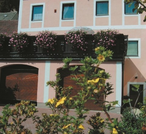 Gästehaus Thaya, © Gästehaus Thaya Ein zweistöckiges rosa Gebäude mit blühenden Pflanzen auf dem Balkon und gelben Blumen im Vordergrund.
