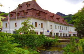 Pension Sun, © Pension Sun Historisches Gebäude mit rotem Dach an einem Bach, umgeben von grüner Vegetation.