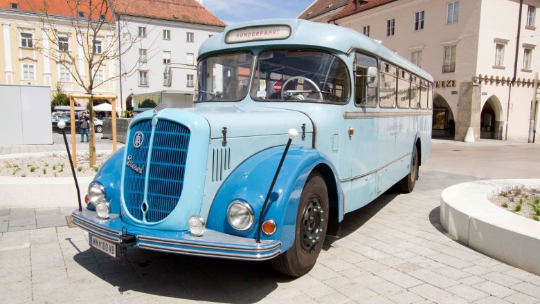 Oldtimer Bus, © Michael Weller Ein blauer Oldtimer-Bus steht auf einem gepflasterten Platz vor historischen Gebäuden.