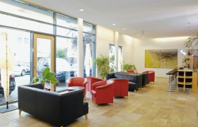 Hotel Klinglhuber, © Steve Haider Helle Hotel-Lobby mit roten und schwarzen Sesseln, Barhockern und großen Fenstern.