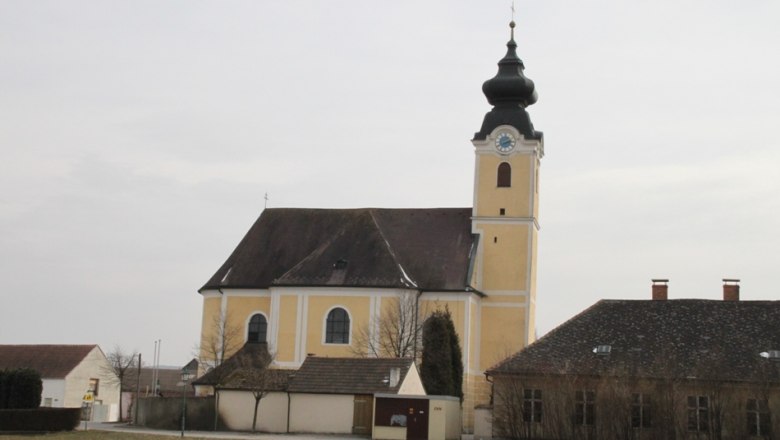 Langenrohr, © Marktgemeinde Langenrohr Gelbe Kirche mit Turm und Uhr in Langenrohr, umgeben von Gebäuden und Bäumen.
