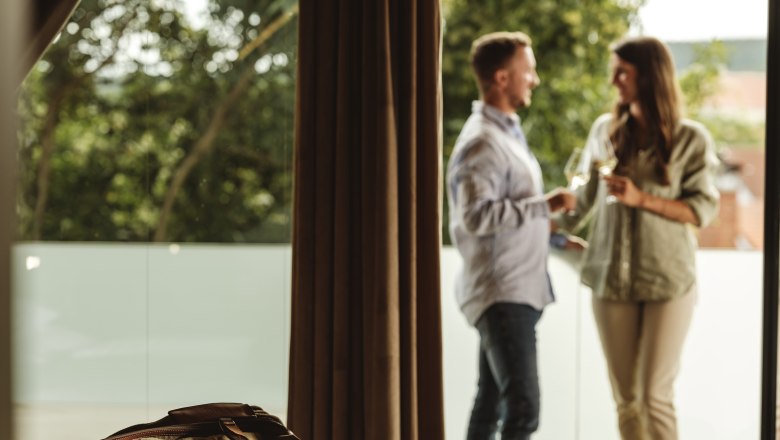 Weingut Hagn Kellerstöckl, © Weinviertel Tourismus GmbH Ein Paar steht auf einem Balkon und hält Weingläser, im Vordergrund liegt eine Reisetasche auf einem Bett.