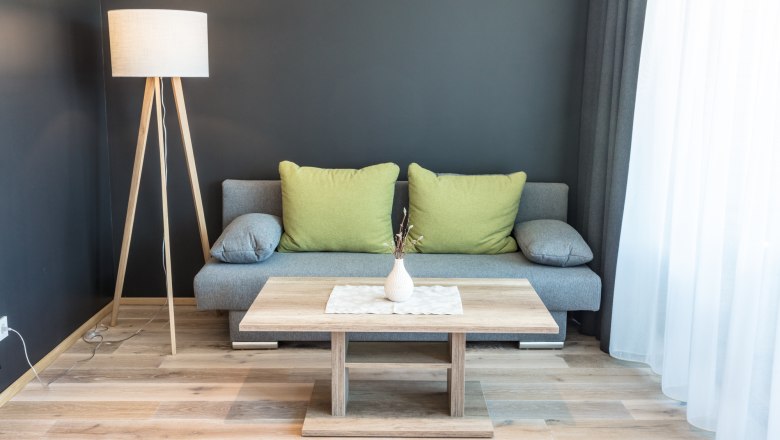 Wohnzimmerbeispiel in der Weinlodge, © KASTNER NEW MEDIA - kastner.tv Modernes Wohnzimmer mit grauem Sofa, grünen Kissen, Holzlampe und Couchtisch vor dunkler Wand.