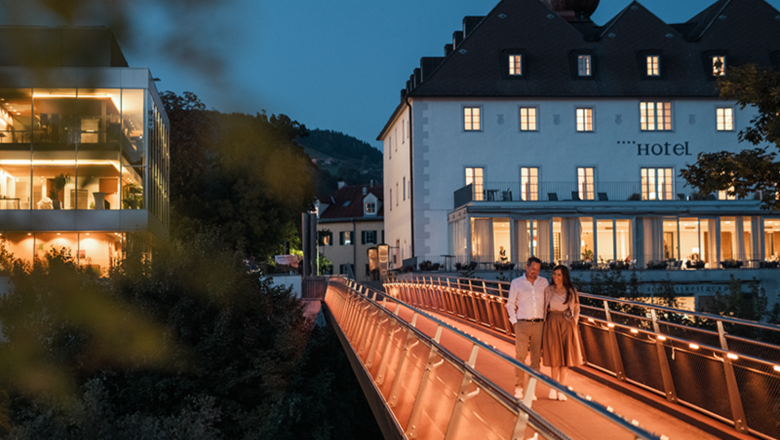 Das Schloss an der Eisenstrasse, © Dominik Stixenberger Ein Paar spaziert abends über eine beleuchtete Brücke vor einem Hotel.