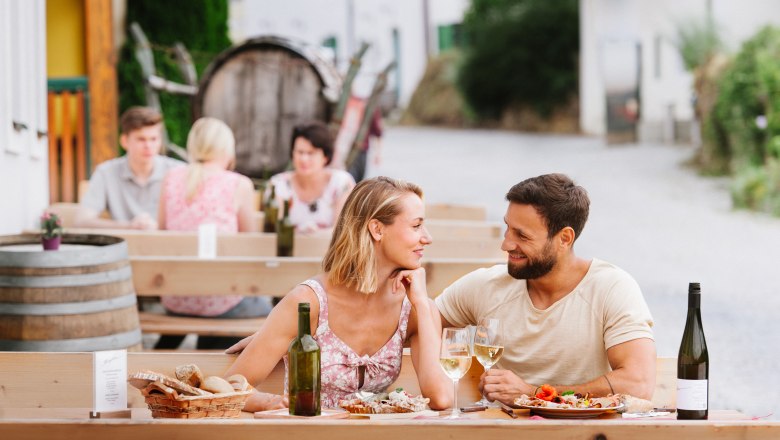 Beim Heurigen in der Kellergasse, © Martina Siebenhandl Ein Paar sitzt an einem Tisch im Freien, genießt Wein und Essen. Im Hintergrund sind weitere Gäste und ein Weinfass zu sehen.