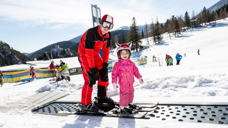 Am Fuße des Schneebergs Schifahren lernen, © NÖ Schneebergbahn, Franz Zwickl Ein Skilehrer hilft einem kleinen Kind beim Skifahren auf einem Förderband im Schnee.