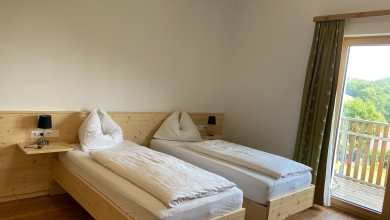 Zweibettzimmer, © Wiener Alpen Zweibettzimmer mit Holzboden, zwei Einzelbetten, Nachttischlampen und einem Balkon mit Ausblick ins Grüne.