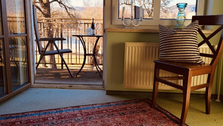 Zimmer im Haupthaus, © Szilágyi Ein Zimmer mit Teppich, Stuhl und Blick auf einen Balkon mit Tisch und Stühlen, Bäume im Hintergrund.