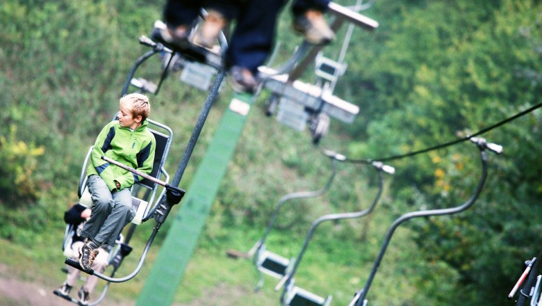 Liftfahrt auf den Muckenkogel, © weinfranz.at Ein Junge in grüner Jacke sitzt auf einem Sessellift, umgeben von grüner Natur.