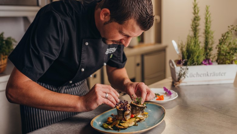 Markus Kraus, © Niederösterreich Werbung/Daniela Führer Ein Koch richtet ein Gericht auf einem Teller an, im Hintergrund sind Küchenkräuter zu sehen.