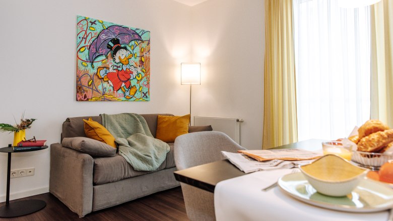 Apartment HEINhotel vienna airport, © HEINhotel vienna airport Wohnzimmer mit grauem Sofa, bunter Comic-Kunst an der Wand, gelben Vorhängen und Frühstückstisch.