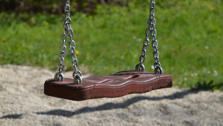 Schaukel, © pixabay Nahaufnahme einer leeren Schaukel aus Holz mit Metallketten, die über einem Kiesboden hängt. Im Hintergrund ist unscharfes Gras zu sehen.