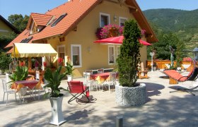 Gästehaus mit Terrasse, Tischen, Stühlen und Sonnenschirmen, umgeben von Pflanzen und Bergen im Hintergrund.
