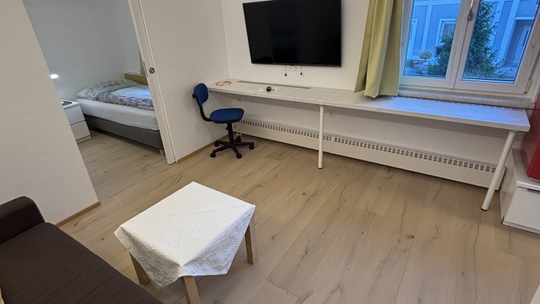 Modernes Wohnzimmer mit braunem Sofa, kleinem Tisch, Fernseher an der Wand, Schreibtisch und Stuhl. Fenster mit Blick auf ein blaues Geb&auml;ude.