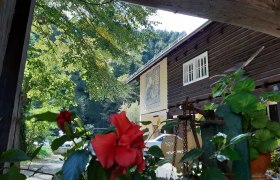 Ein traditionelles Holzhaus mit roten Blumen im Vordergrund und gr&uuml;nen B&auml;umen im Hintergrund.