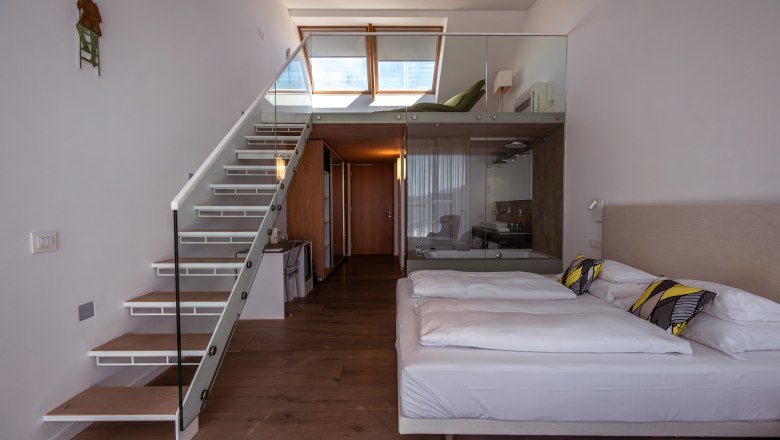 Modernes Hotelzimmer mit Treppe zu einem Zwischengeschoss, Doppelbett und Glaswand zum Badezimmer.