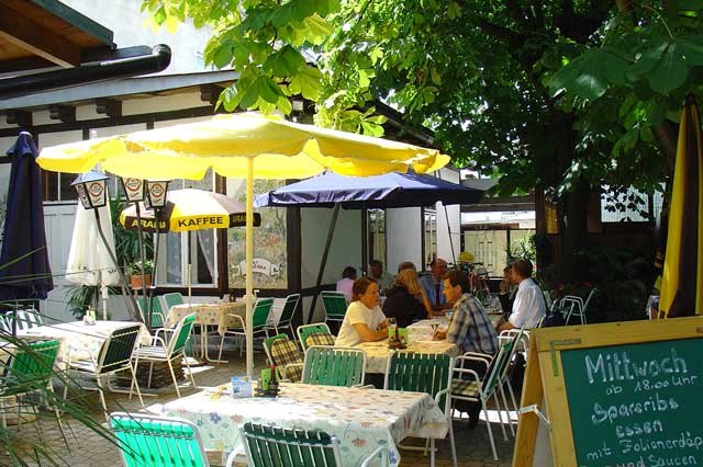 Ein gem&uuml;tlicher Gastgarten mit Tischen, St&uuml;hlen und Sonnenschirmen, umgeben von B&auml;umen. Menschen sitzen und unterhalten sich.