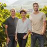 Drei Personen stehen in einem Weinberg, umgeben von grünen Reben, mit einem Dorf im Hintergrund.