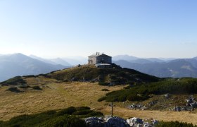 Berghütte auf einem Hügel mit Bergpanorama im Hintergrund.