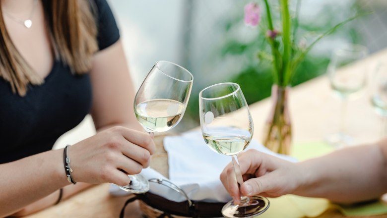 Zwei Personen stoßen mit Weißweingläsern an einem Tisch an.