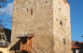 Ein alter Wehrturm mit h&ouml;lzerner Treppe und Gel&auml;nder, umgeben von Geb&auml;uden, unter blauem Himmel.