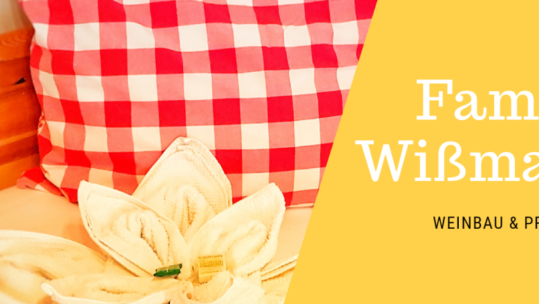 Herzlich Willkommen!, © Fam. Wißmann Rotes Kissen mit Karomuster und Handtuch in Blumenform auf einem Bett. Text: Familie Wißmann, Weinbau & Privatzimmer.