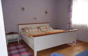 Ein Schlafzimmer mit einem weißen Bett, bunten Bettdecken, einem Nachttisch und einem Fenster mit Vorhängen.