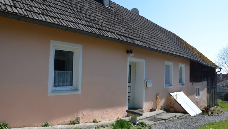 Ein einfaches, einst&ouml;ckiges Haus mit rosa Fassade und grauem Dach. Vor dem Haus sind Pflanzen und Dekorationen zu sehen.