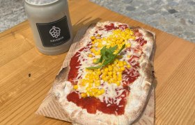 Fladen mit Mais und Rucola auf einem Holzbrett serviert, daneben ein Behälter mit Besteck und Servietten.