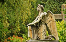 Engelsstatue auf einem Friedhof, umgeben von gr&uuml;ner Vegetation.