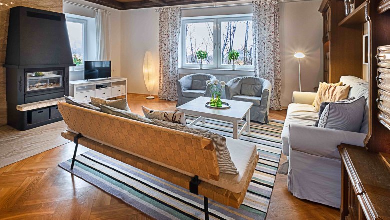 Top 1 - Wohnzimmer, © VP FeWo OG Gemütliches Wohnzimmer mit Kamin, Sofa, Sessel und Fernseher. Holzboden und Decke, gestreifter Teppich, große Fenster mit Vorhängen.