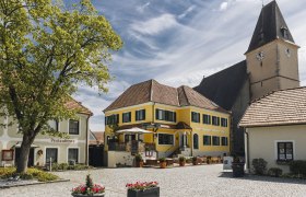 Idyllischer Dorfplatz mit Gasthaus und Terrasse und ein Kirchturm im Hintergrund.