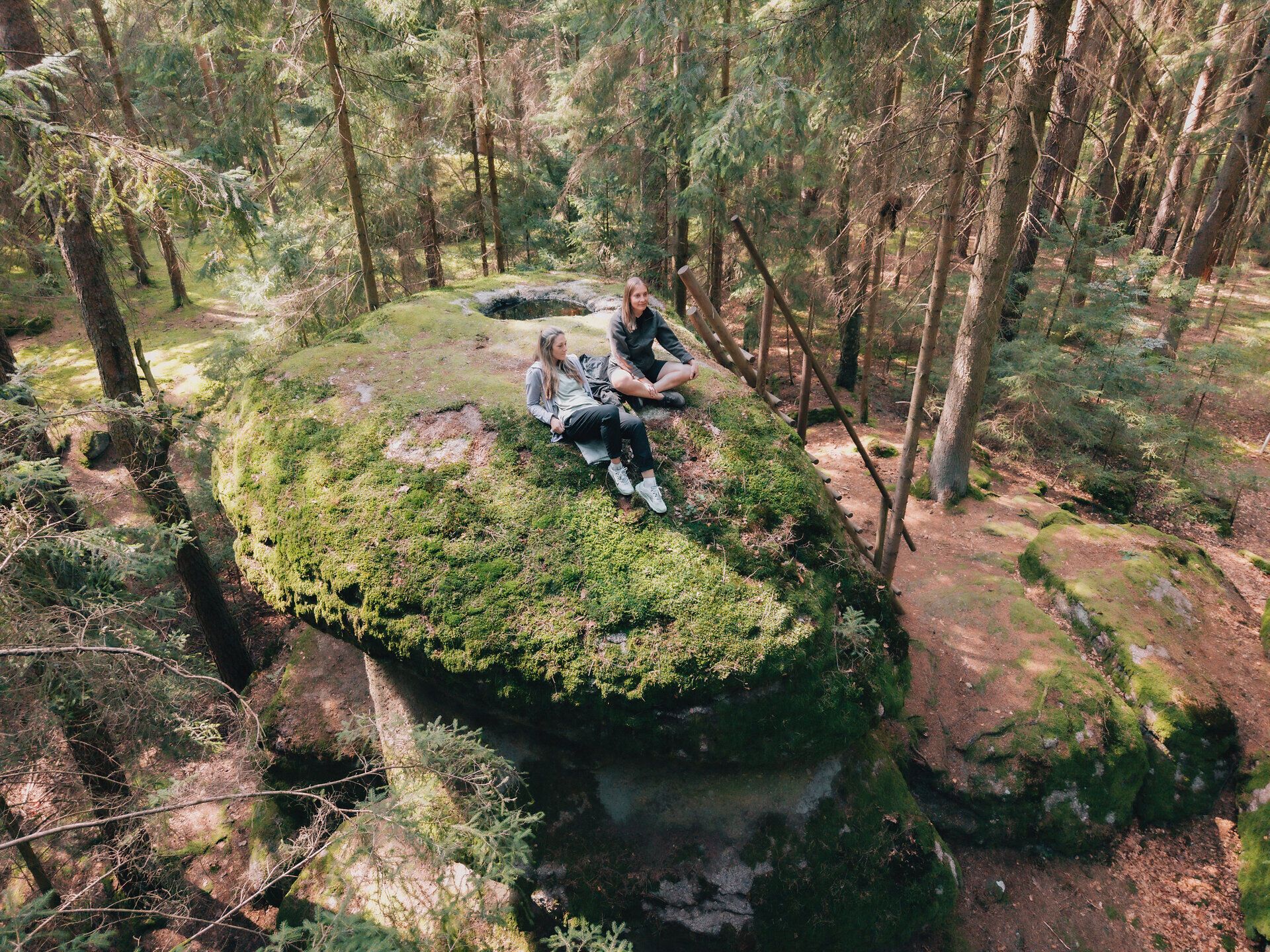 Zwei Frauen sitzen entspannt auf dem moosbewachsenen Felsen Kierlingstein in der Kraftarena Groß Gerungs im Waldviertel, umgeben von dichtem Wald und hohen Nadelbäumen.