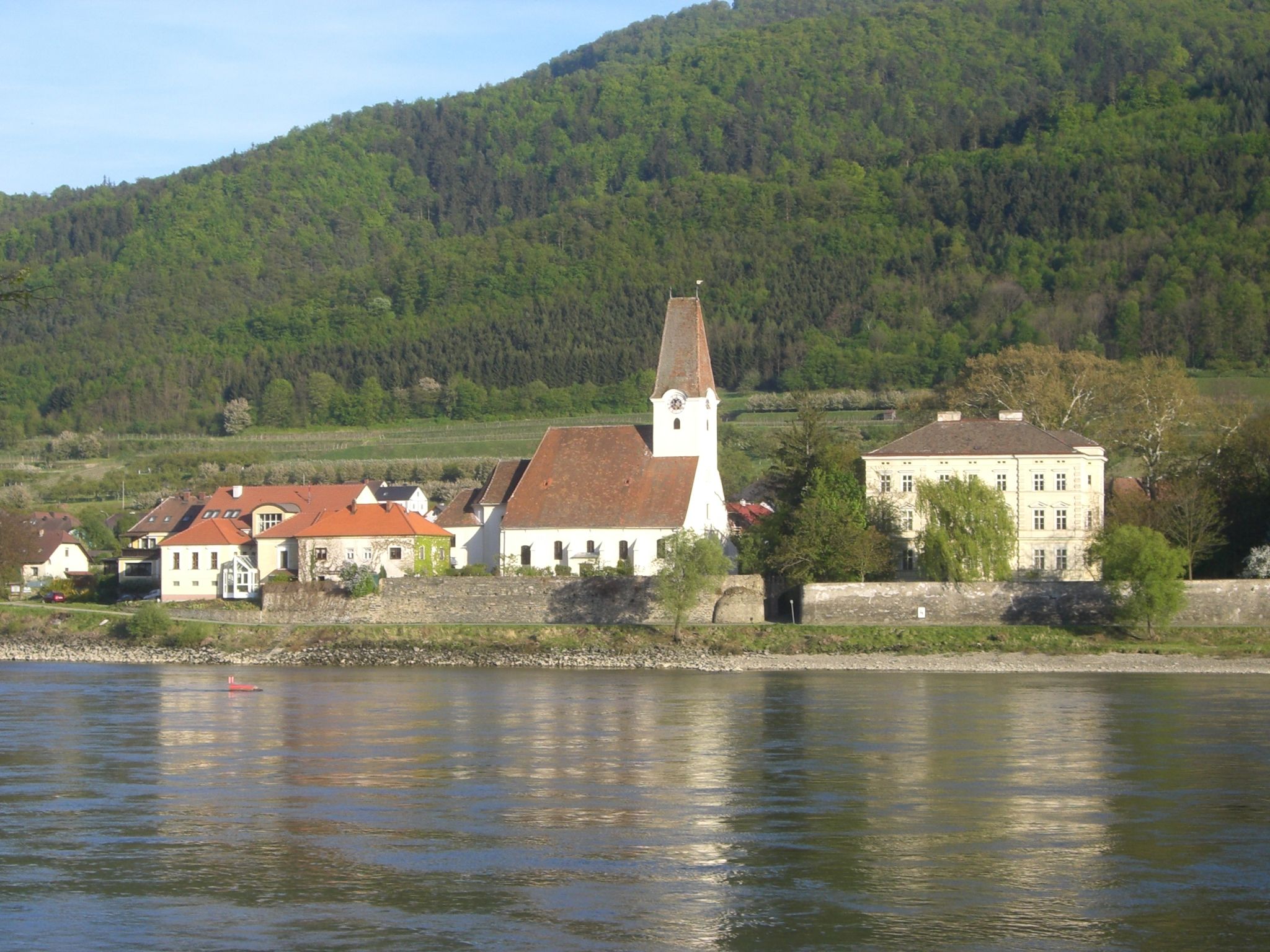 Pfarrkirche Hofarnsdorf mit umliegenden Gebäuden und bewaldetem Hügel im Hintergrund, aufgenommen von der Donau aus.