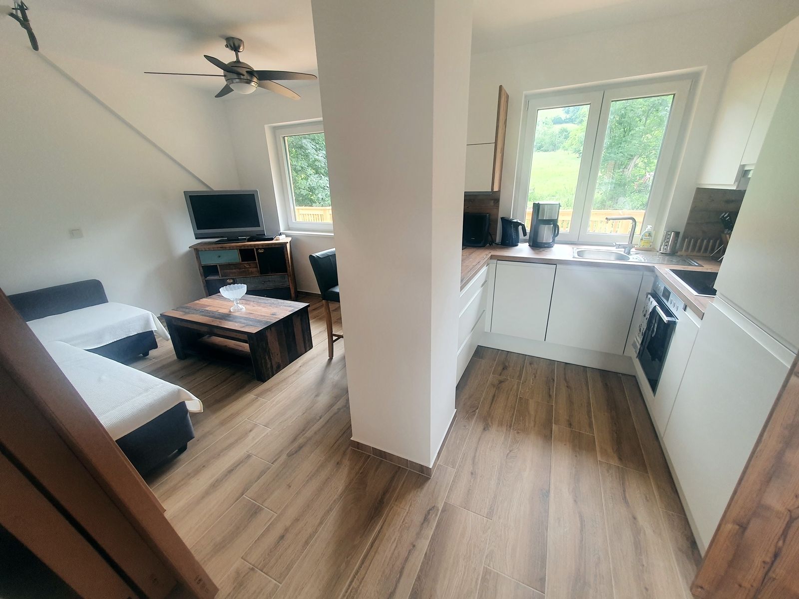 Moderne Wohnküche mit Holzfußboden, Sofa, Fernseher, Couchtisch und weißer Einbauküche mit Fenster.
