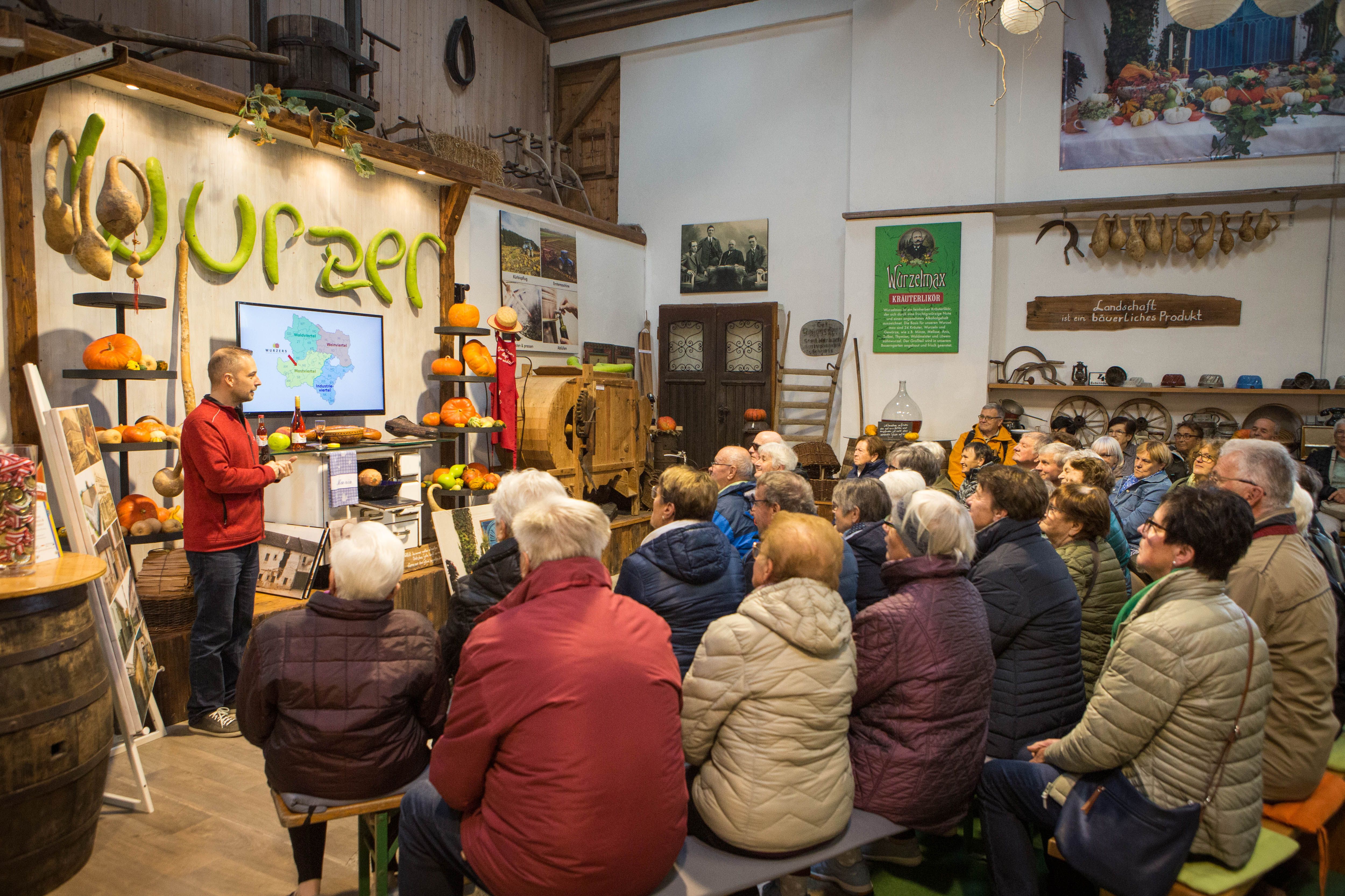 Ein Mann hält einen Vortrag vor einer Gruppe von Menschen in einem rustikalen Raum mit Kürbissen und landwirtschaftlichen Werkzeugen.