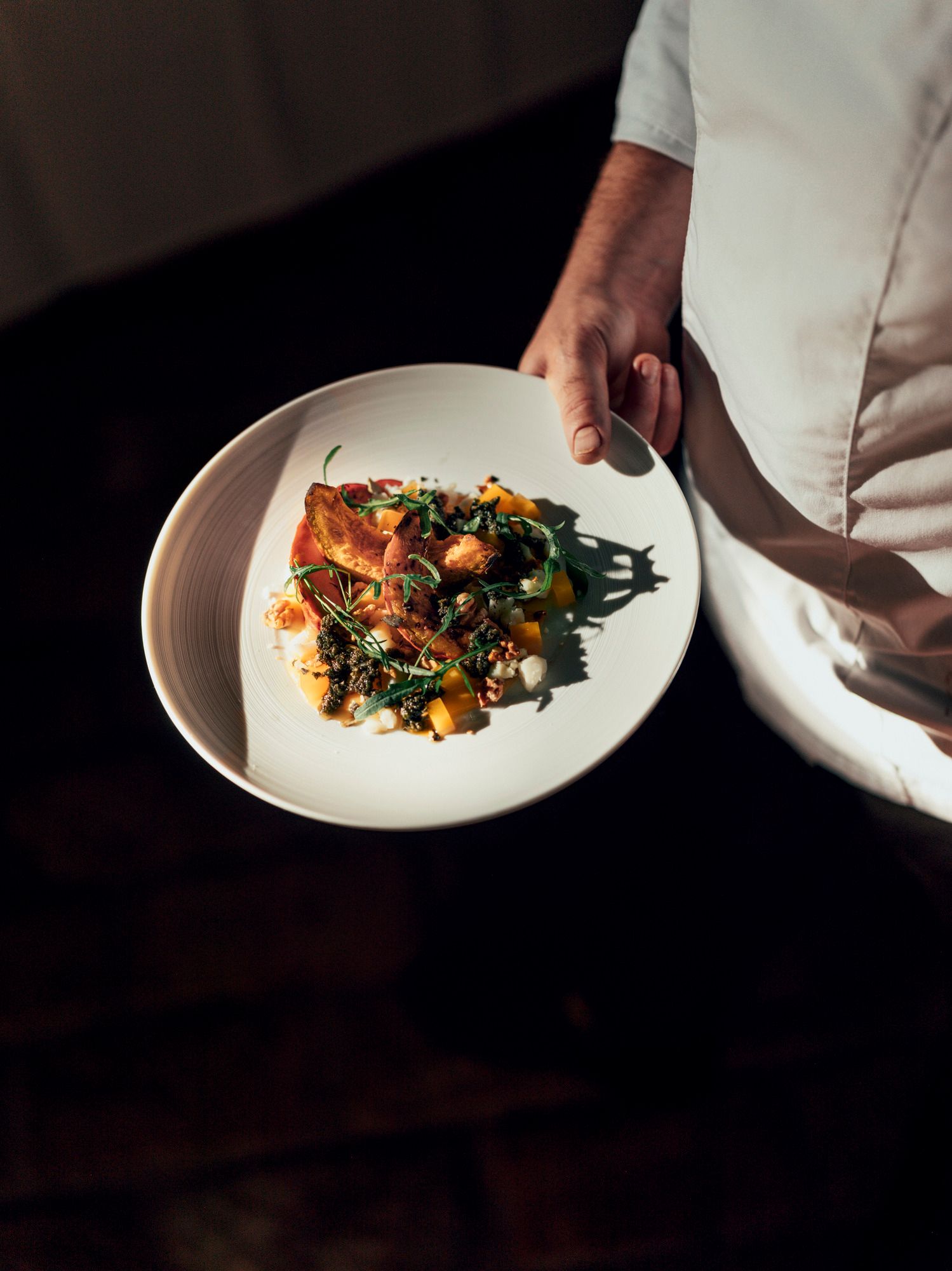 Ein Teller mit geröstetem Hokkaido-Kürbis, Salbeipesto und Schafkäse, gehalten von einer Person in weißer Kleidung.
