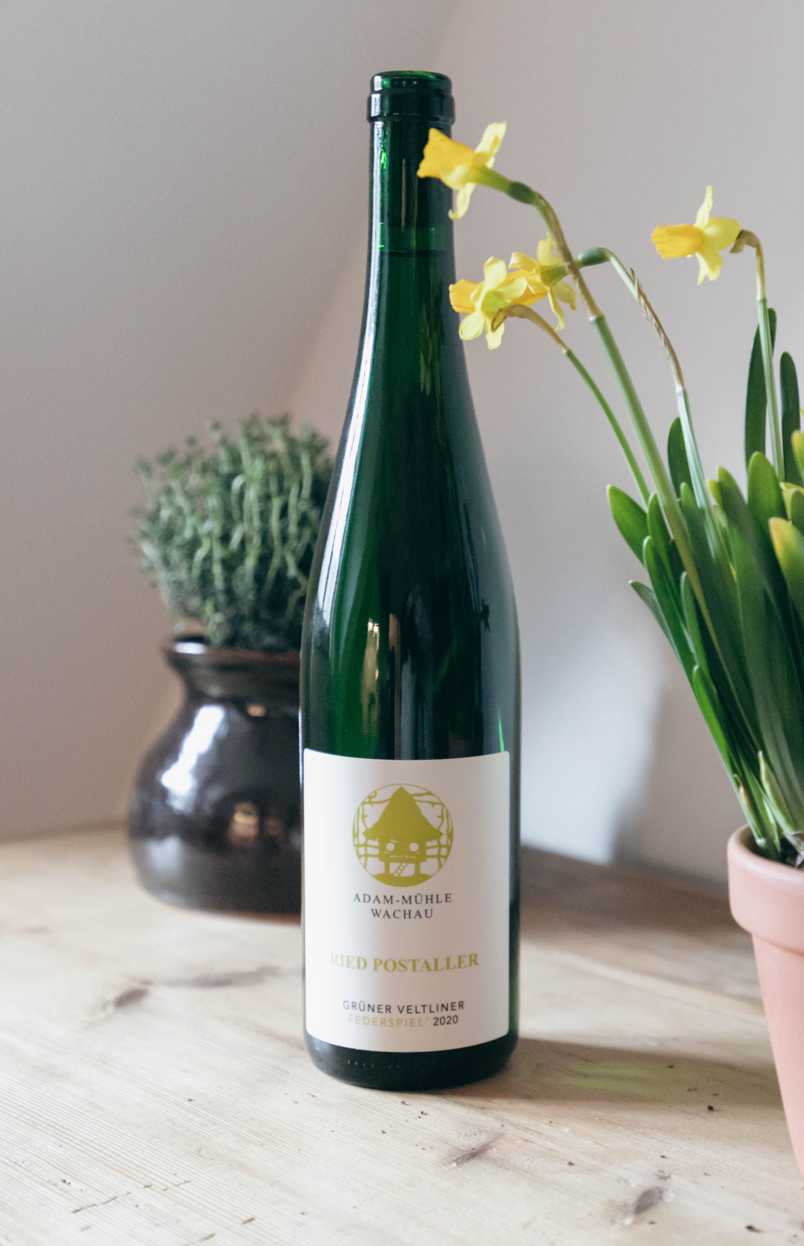 Eine Flasche Grüner Veltliner Wein steht auf einem Tisch neben gelben Blumen und einem Topf mit Kräutern.