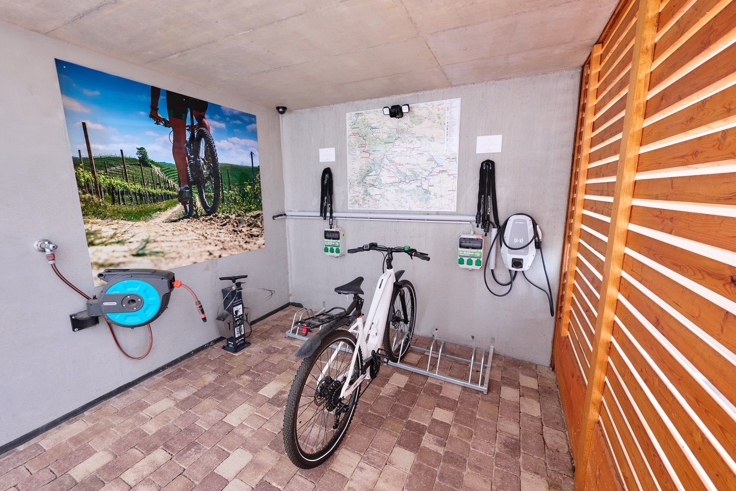 Fahrrad in einer Garage mit Ladestation und Wandbild eines Radfahrers in einem Weinberg.
