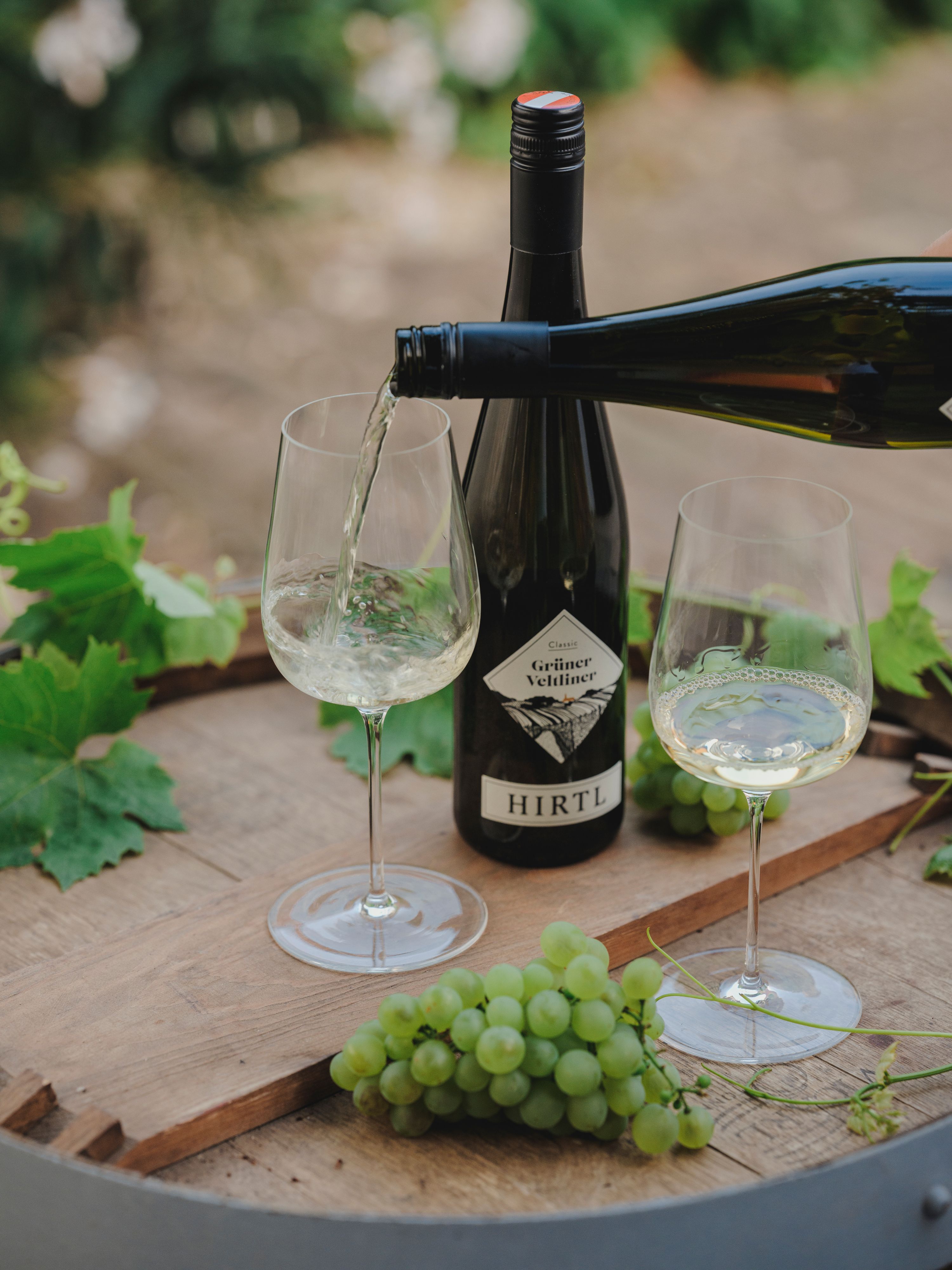 Weißwein wird in ein Glas gegossen, daneben eine Flasche Grüner Veltliner von Hirtl und grüne Trauben auf einem Holztisch.