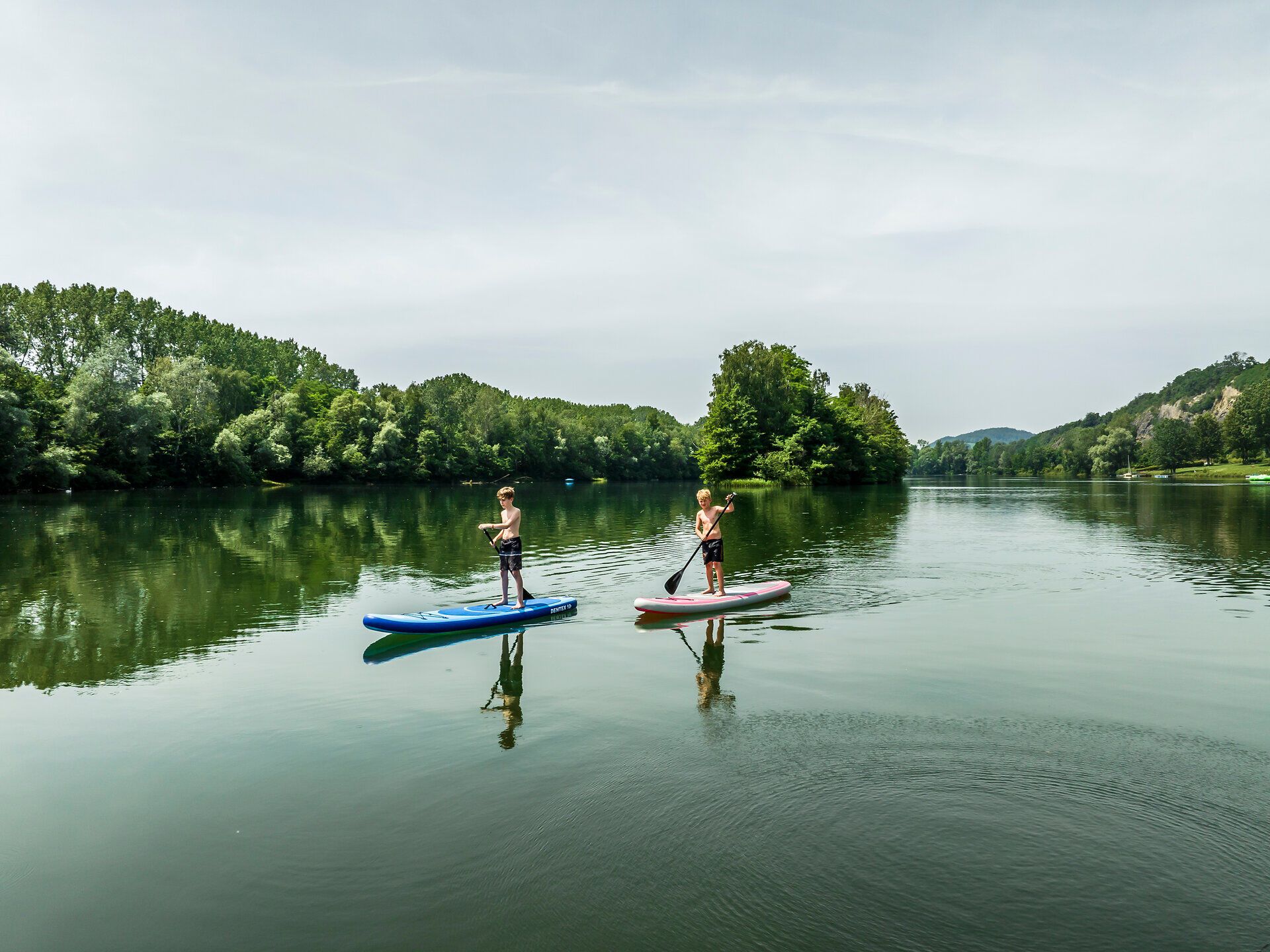 Drei Personen auf einem Tretboot am See, Fahrräder am Steg, grüne Bäume im Hintergrund.