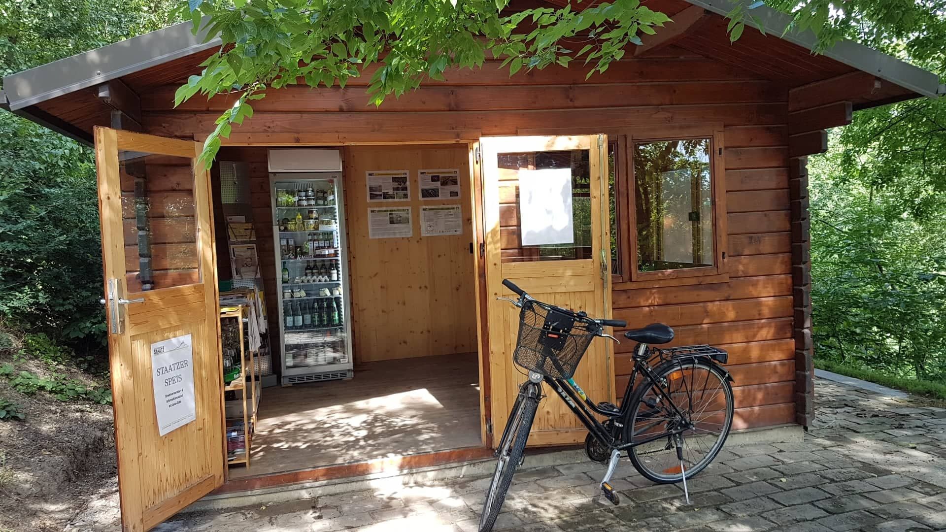 Kleines Holzhaus mit offenem Eingang, Kühlschrank mit Getränken und einem Fahrrad davor.