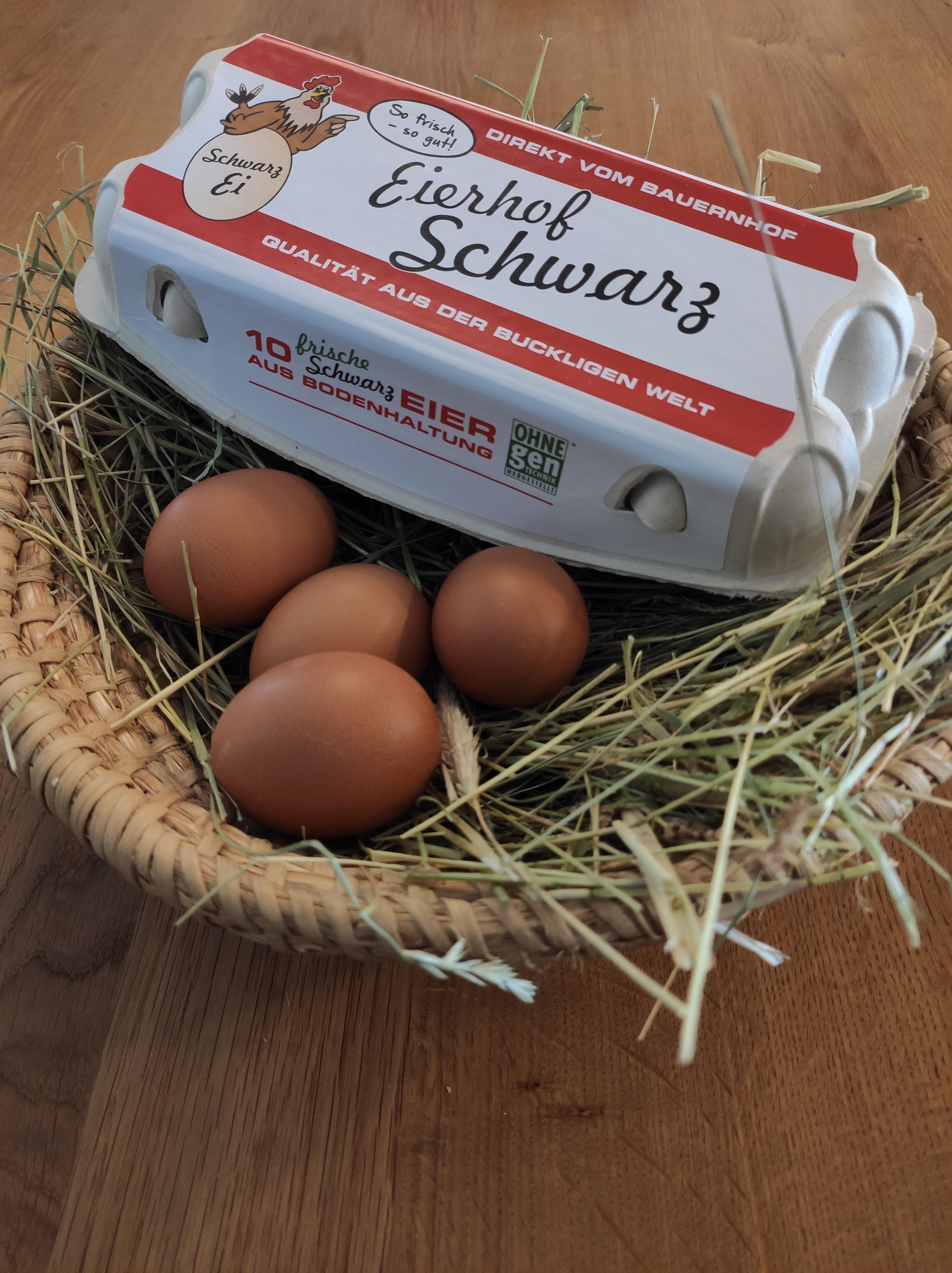 Ein Eierkarton mit der Aufschrift 'Eierhof Schwarz' liegt auf einem Korb mit Stroh und braunen Eiern.