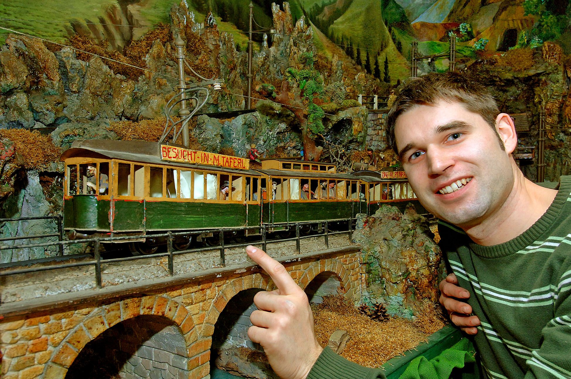 Ein Mann zeigt auf eine Modelleisenbahn in einer alpinen Landschaft.