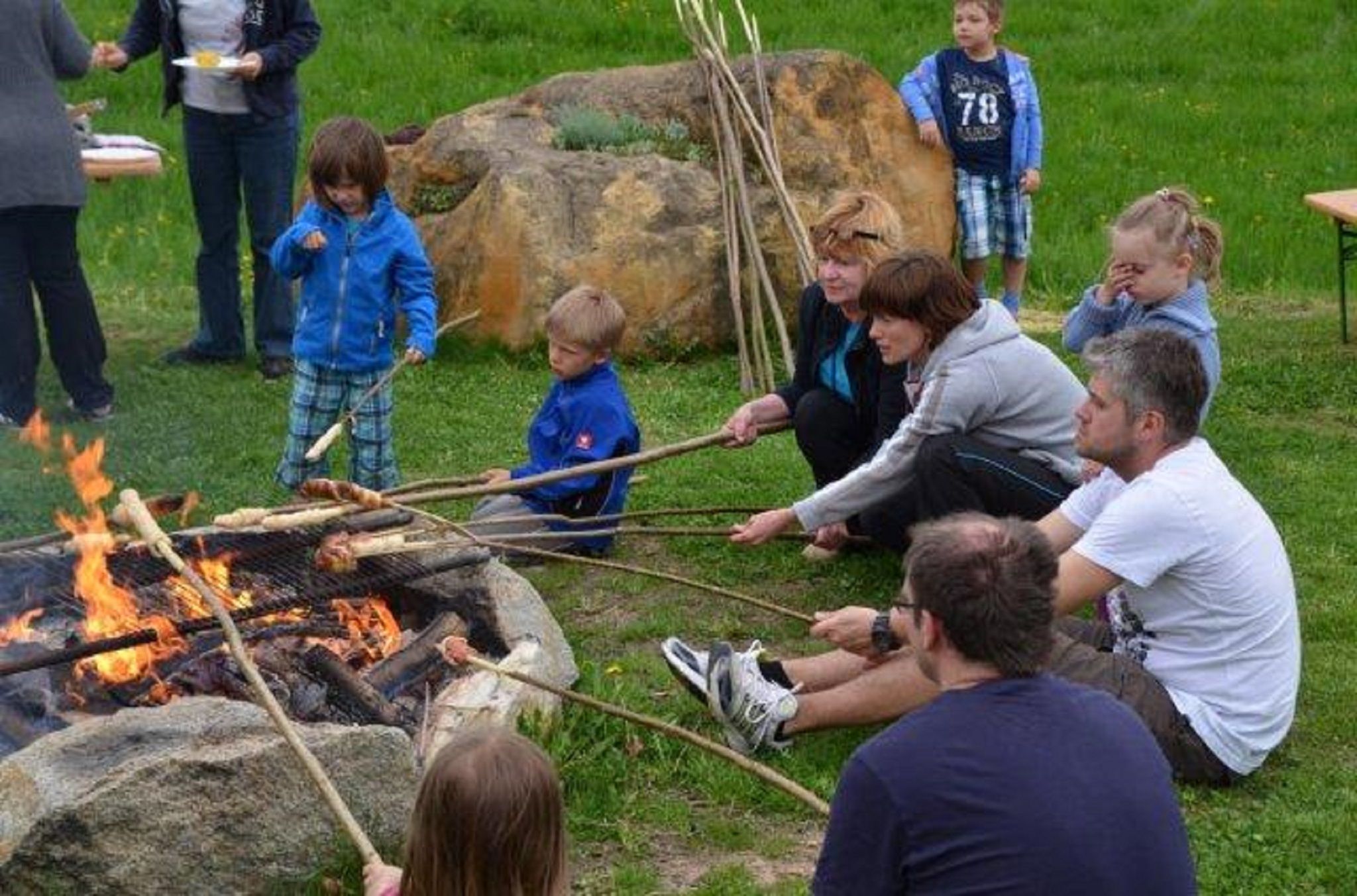 Menschen sitzen um ein Lagerfeuer und rösten Brot an langen Stöcken.