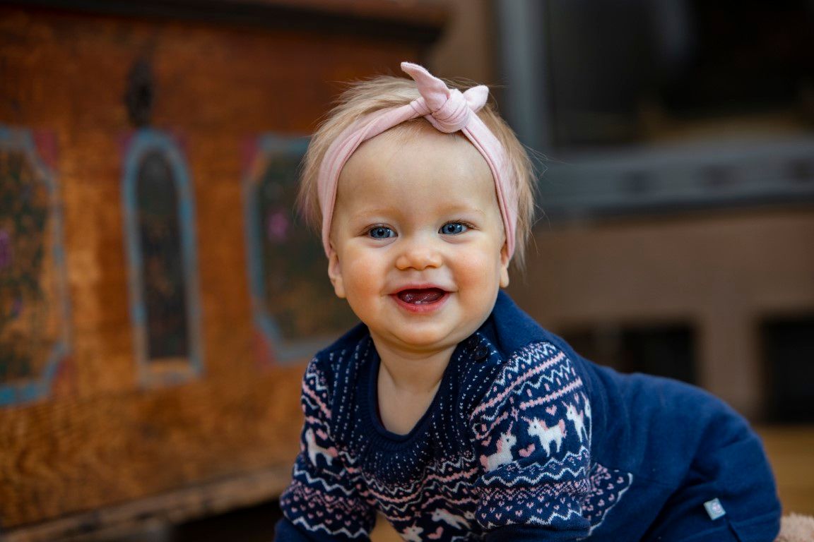 Ein lächelndes Baby mit rosa Stirnband und blauem Pullover mit Rentiermuster.