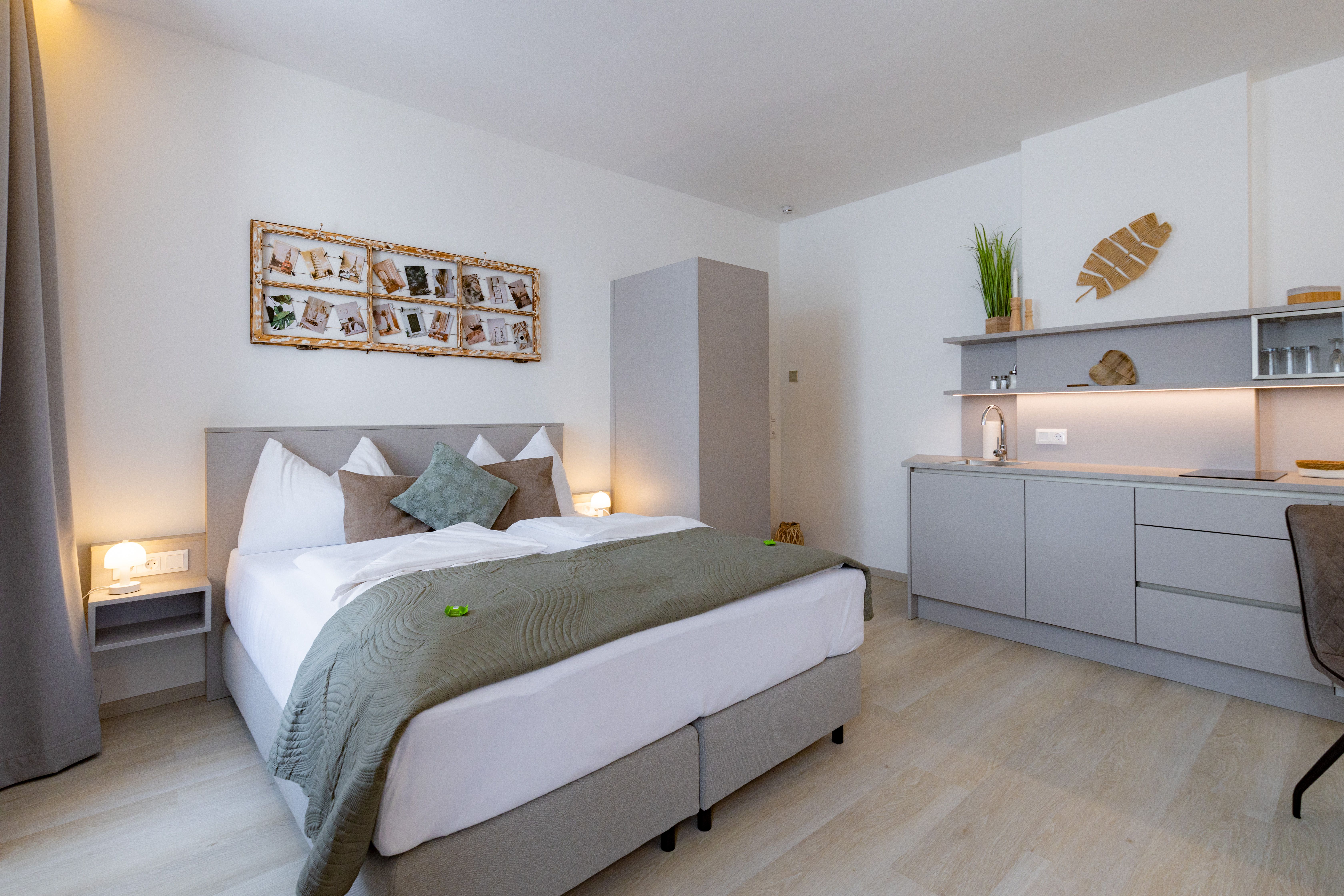 Modernes Schlafzimmer mit Doppelbett, grauer Bettwäsche, Wanddekoration und kleiner Küchenzeile.